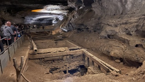A dimly lit cave interior with wooden structures and excavation sites, surrounded by rocky walls and visitors in the background.
