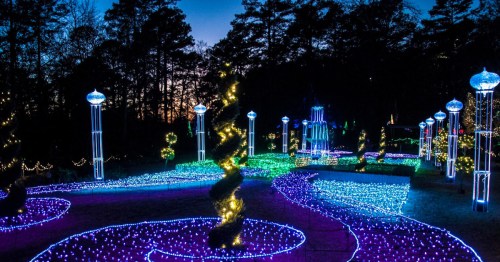 A colorful display of illuminated pathways and trees in a park at dusk, surrounded by glowing lights.