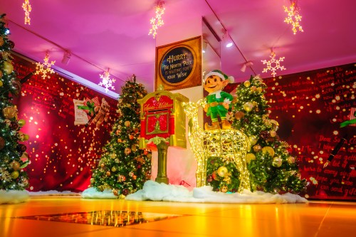A festive scene with decorated Christmas trees, a glowing mailbox, and an elf figure in a colorful holiday setting.