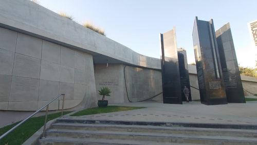 A modern memorial with sleek black structures and a curved concrete wall, surrounded by greenery and steps.