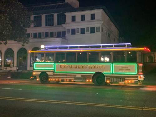 A brightly lit trolley bus parked at night, with colorful neon lights and a building in the background.
