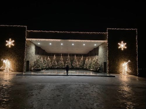 A festive outdoor scene at night, featuring decorated Christmas trees and lights on a stone building.