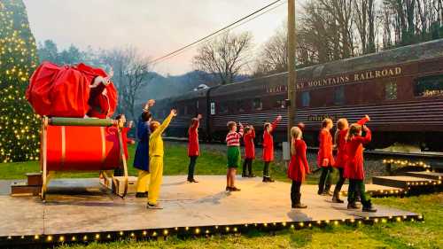 Children in festive costumes perform on stage near a decorated train, with a large Santa sleigh prop and holiday lights.