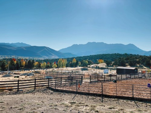 Scenic view of a ranch with mountains in the background, colorful trees, and a clear blue sky.