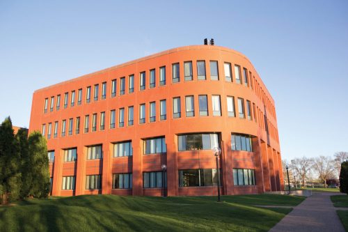 A modern red brick building with large windows, surrounded by green grass and trees under a clear blue sky.