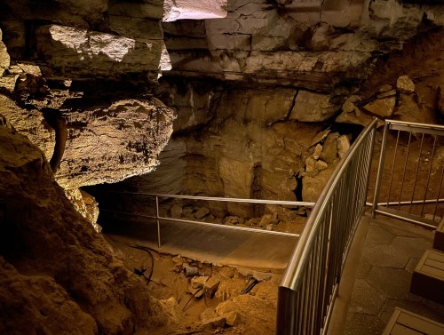 A dimly lit cave interior with rocky walls and a metal railing along a pathway leading into the depths.