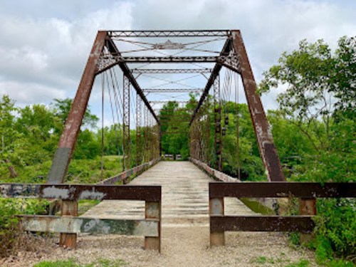 Rusty metal bridge spanning a river, surrounded by lush green trees and a cloudy sky.