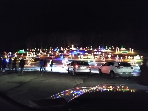 A festive display of colorful holiday lights illuminates a park, with people admiring the decorations from parked cars.