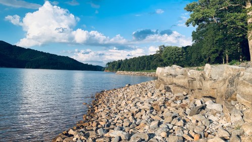 A serene lakeside view with rocky shorelines, lush green trees, and a blue sky dotted with clouds.