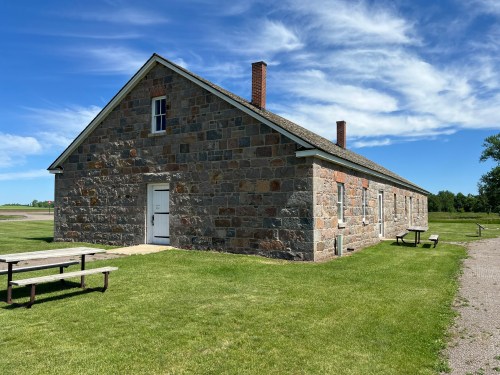 A stone building with a gabled roof, surrounded by green grass and picnic tables under a blue sky with clouds.