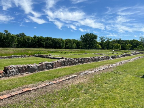 Ruins of stone foundations in a grassy field under a blue sky with scattered clouds.