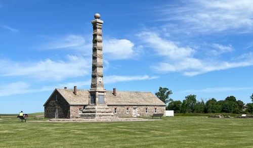 A tall stone monument stands beside a historic building on a grassy field under a clear blue sky.