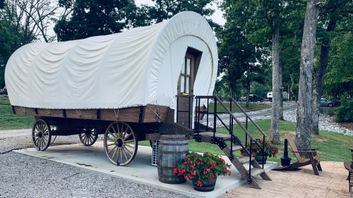 A covered wagon on a concrete pad, surrounded by flowers and trees, with a gravel path nearby.