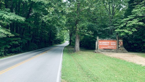 A scenic road leads to the entrance of Mammoth Cave National Park, surrounded by lush green trees.