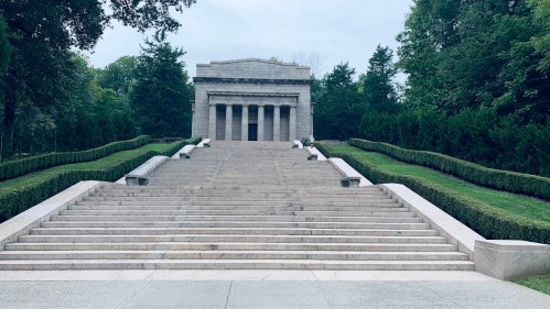 A grand stone monument with steps leading up, surrounded by greenery and trees under a cloudy sky.