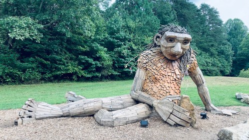 A large wooden sculpture of a seated giant with a friendly expression, surrounded by greenery.
