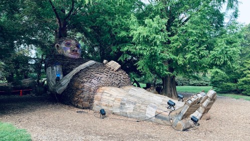 A large wooden sculpture of a reclining figure surrounded by trees, with lights illuminating its features.
