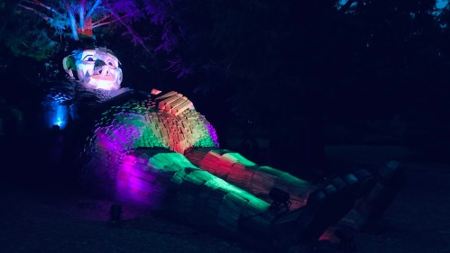 A large, colorful wooden sculpture of a reclining figure illuminated by vibrant lights in a dark outdoor setting.