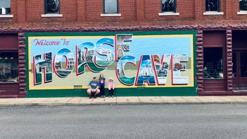 Two children and an adult pose in front of a colorful mural that reads "Welcome to Horse Cave."