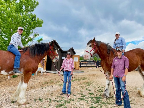 Two people on horses and two standing beside them, all in a rural setting with a cloudy sky.