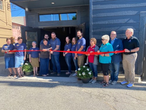 A group of people stands in front of a building, holding a red ribbon for a ribbon-cutting ceremony.