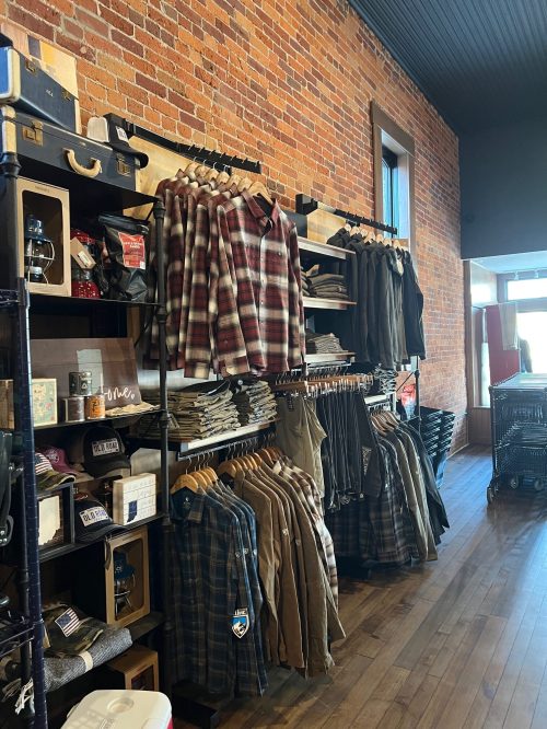 A clothing store interior featuring shelves of flannel shirts and pants against a brick wall.