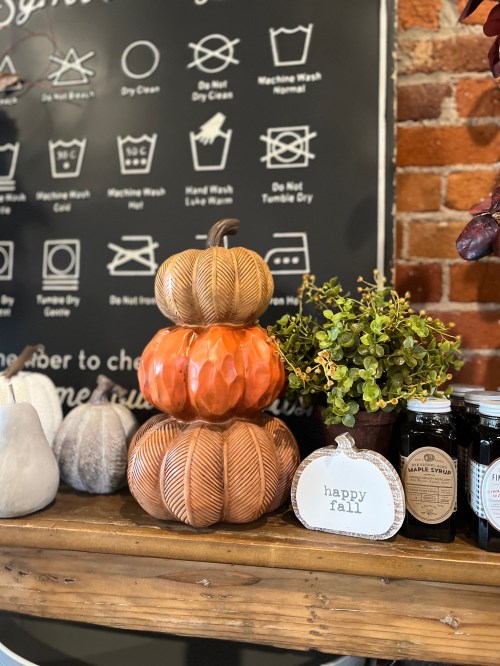 A stack of three decorative pumpkins in autumn colors, with a small plant and a "happy fall" sign on a wooden shelf.