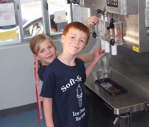 Two children at an ice cream shop, one filling a cup with soft serve while the other smiles playfully behind.