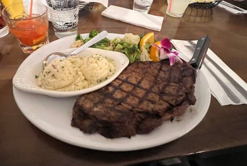 A plate with a grilled steak, garlic mashed potatoes, and vegetables, with drinks and condiments on the table.