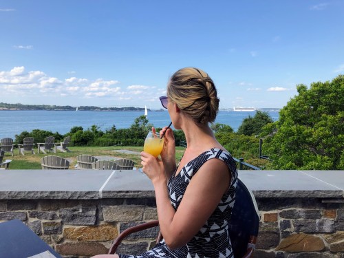 A woman in a patterned dress enjoys a drink while overlooking a scenic waterfront view with sailboats.