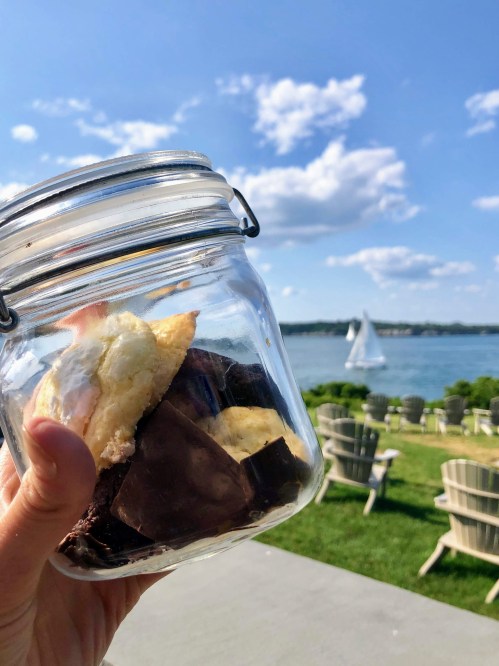 A hand holding a jar of baked goods with a scenic waterfront and sailboat in the background.