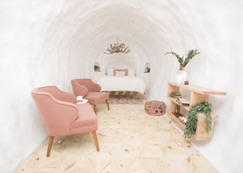 Cozy, minimalist room with a curved white wall, two pink chairs, a bed, and wooden decor elements.