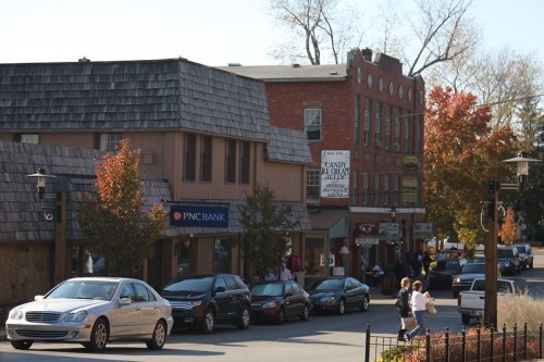 A street scene featuring shops, trees with autumn leaves, and parked cars in a small town.