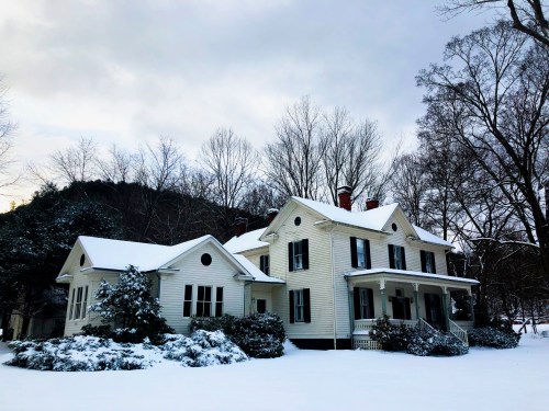 A large, white house surrounded by snow, with trees and mountains in the background under a cloudy sky.