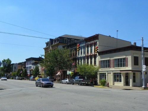 A sunny street scene featuring historic buildings, trees, and cars on a quiet road. Traffic lights are visible.