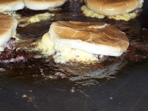 Cheeseburgers cooking on a griddle, with melted cheese oozing out from between the buns.