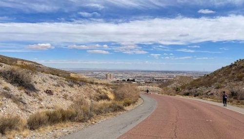 A winding road descends through a hilly landscape, with a city skyline visible in the distance under a blue sky.