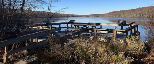 A wooden dock overlooking a calm lake surrounded by trees, with clear blue skies and autumn foliage in the background.