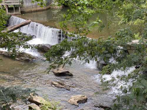 A serene waterfall cascades over rocks, surrounded by lush greenery and a calm river.