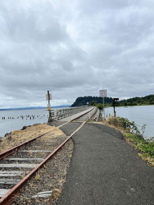 A narrow path alongside a railway track leads to a bridge over calm water, with cloudy skies in the background.