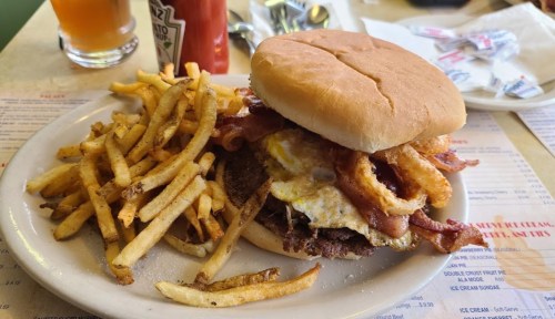 A hearty burger topped with bacon, fried egg, and onion rings, served with a side of crispy French fries.