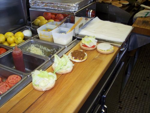 A countertop with burger buns, lettuce, tomatoes, and condiments in a kitchen setting.