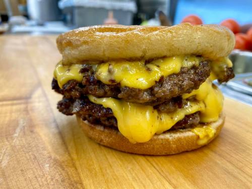 A stacked cheeseburger with three beef patties and melted cheese, served on a toasted bun, on a wooden countertop.