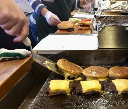A chef flips burgers with melted cheese on a grill, while assembling sandwiches in the background.