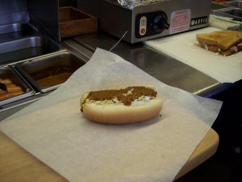 A hot dog in a bun topped with a brown sauce, placed on parchment paper in a kitchen setting.