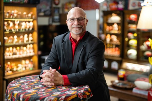 A smiling man in a black suit and red shirt sits at a table, surrounded by sports memorabilia in a cozy room.