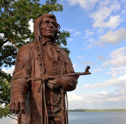 A bronze statue of a Native American figure stands by a lake, surrounded by trees and a blue sky.