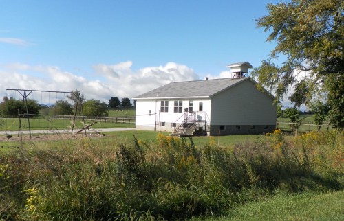 A rural schoolhouse with a playground in front, surrounded by green fields and a blue sky with clouds.