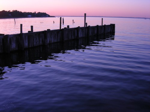 A serene waterfront scene at dusk, featuring a wooden dock and calm water reflecting soft purple and pink hues.