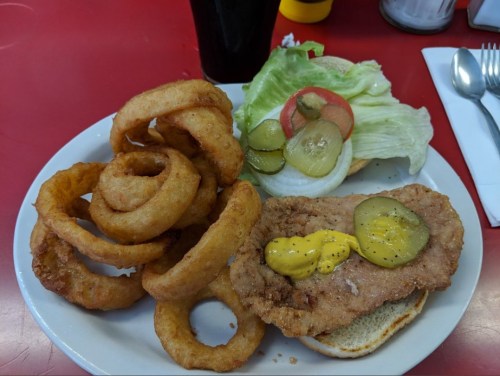 A plate with a fried chicken sandwich topped with pickles and mustard, served with onion rings and a side of lettuce and tomato.